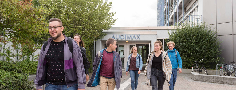 Sechs Studierende laufen teilweise zueinander gewandt aus dem Audimaxgebäude heraus.